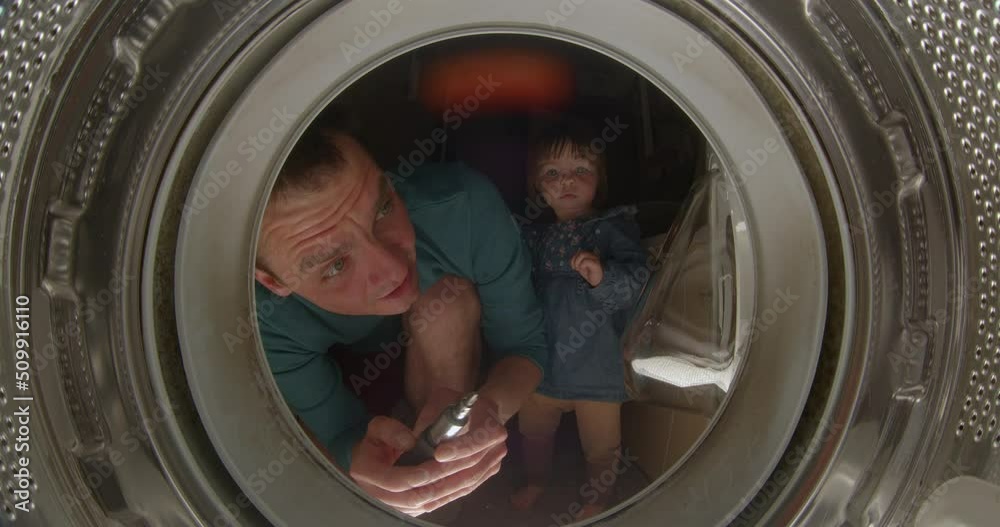 Male father makes repairs to the washing machine. He turns the trust screws with a screwdriver. Nearby is a girl daughter. View from the washing machine, drum. Male father makes repairs to the washing machine. He turns the trust screws with a screwdriver. Nearby is a girl daughter. View from the washing machine, drum.