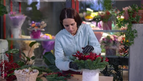 Skilled absorbed Caucasian florist inserting red leaves in bouquet base standing at table in workshop. Portrait of concentrated confident woman creating composition in flower shop indoors