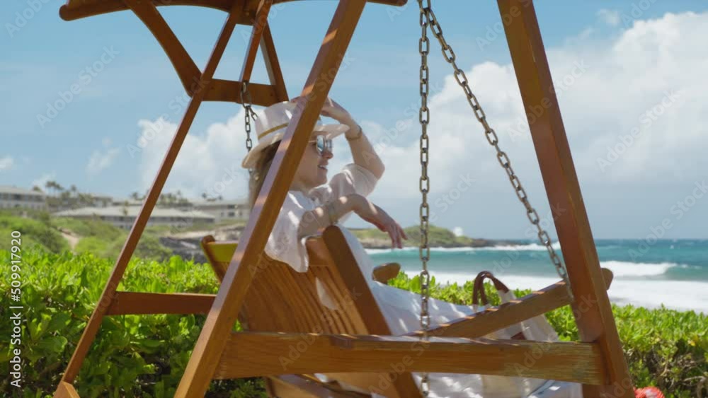 Happy young woman in fashionable white fedora hat with tropical frangipani plumeria flowers on wooden swing at ocean park. Smiling lady sitting on wooden swing and looking at blue ocean on Hawaii 6K Happy young woman in fashionable white fedora hat with tropical frangipani plumeria flowers on wooden swing at ocean park. Smiling lady sitting on wooden swing and looking at blue ocean on Hawaii 6K
