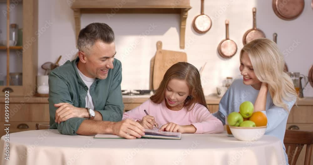 Happy loving parents and cute daughter sitting at kitchen table and doing school exercises together at home. Caucasian mother and father assisting kid with homework. Happy loving parents and cute daughter sitting at kitchen table and doing school exercises together at home. Caucasian mother and father assisting kid with homework.