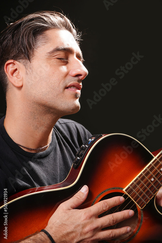 Young man with closed eyes feeling the sounds. holding an electroacoustic guitar, with a black t-shirt and black background. photo