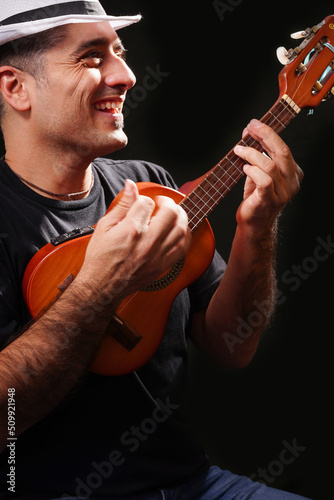 Young man playing happy cavaquinho, with a white hat, black t-shirt and black background. photo
