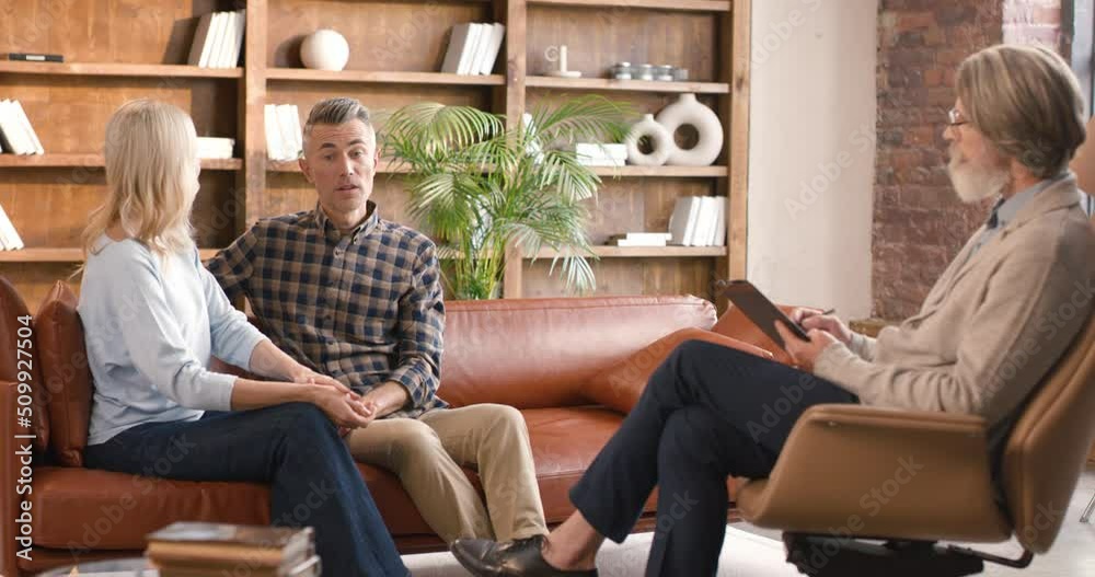Mature attractive woman and handsome man discussing family life with senior bearded therapist during couple consultation in office. Concept of relationship and psychology. Mature attractive woman and handsome man discussing family life with senior bearded therapist during couple consultation in office. Concept of relationship and psychology.