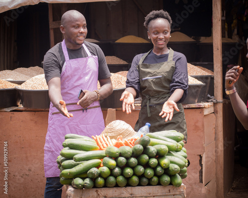 A male and female African Nigerian traders, sellers, business persons or shop owners, having aprons on and happily pointing and attracting potential customers to their displayed goods in a market
