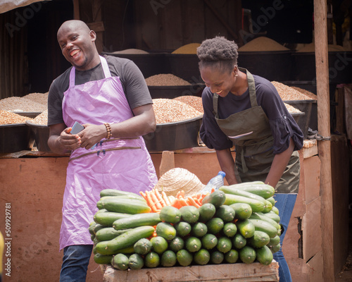 A male and female African Nigerian traders, sellers, business persons or shop owners, having aprons on and laughing together as they wait for potential customers in a market