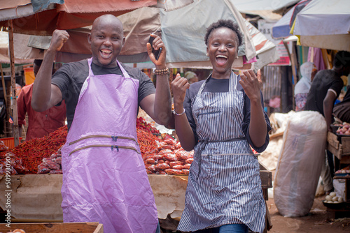 Male and female African Nigerian traders, sellers, business persons or entrepreneurs with aprons, celebrating together at their stall in a market