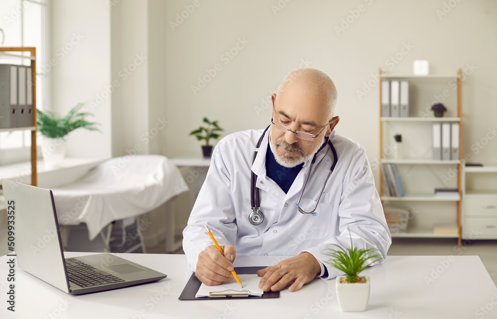 Senior man doctor in glasses sit at desk write in patient medical card work on computer in clinic. Older GP in white uniform prescribe meds consult client online on laptop. Healthcare concept.