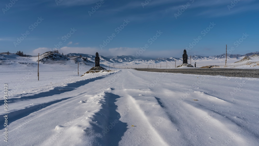 A straight asphalt highway runs through a snow-covered valley. Statues of idols in snowdrifts on the roadsides. Ahead, against the blue sky, a mountain range could be seen. Altai. Chuysky tract