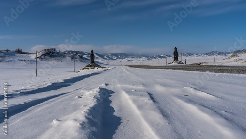 A straight asphalt highway runs through a snow-covered valley. Statues of idols in snowdrifts on the roadsides. Ahead, against the blue sky, a mountain range could be seen. Altai. Chuysky tract