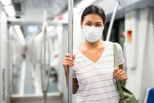 Black-haired asian woman in mask standing inside subway car and holding handrail.