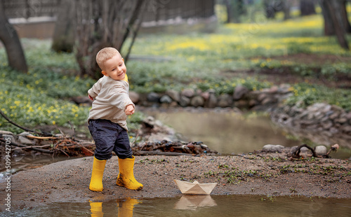 Portrait of adorable little kid boy in rubber boots launch paper boats in a puddle in spring. Origami.