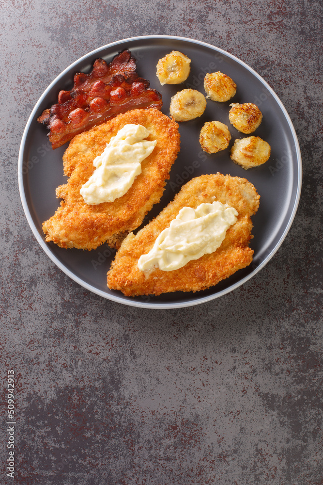 Chicken Maryland is a breaded, pan-fried chicken garnished with bananas and bacon close-up in a plate on the table. Vertical top view from above