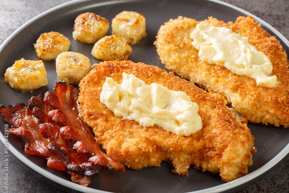 Fried breaded chicken fillet with white sauce served with bananas and bacon close-up on a plate on the table. horizontal