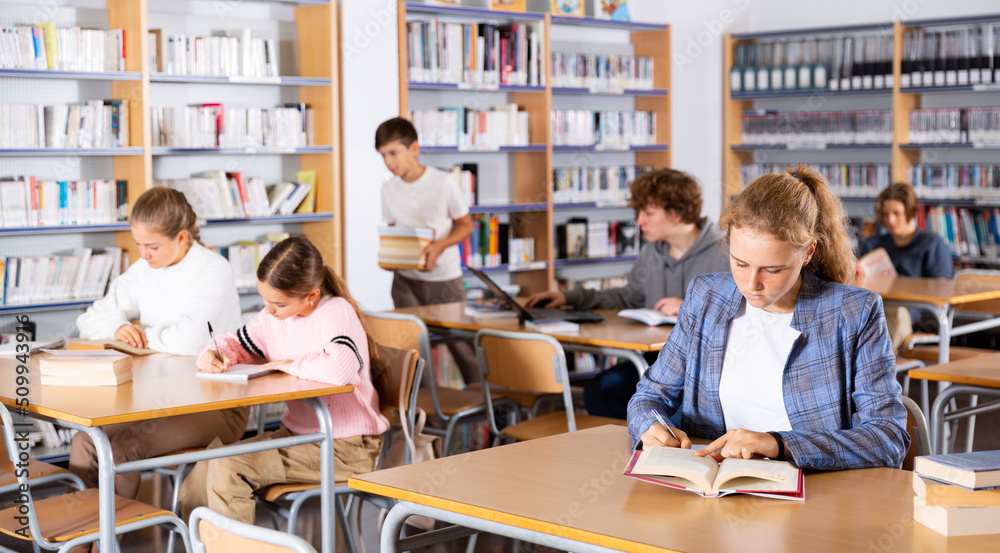 Fifteen-year-old girl, sitting at a desk in the library, writes out an abstract from a textbook in a notebook