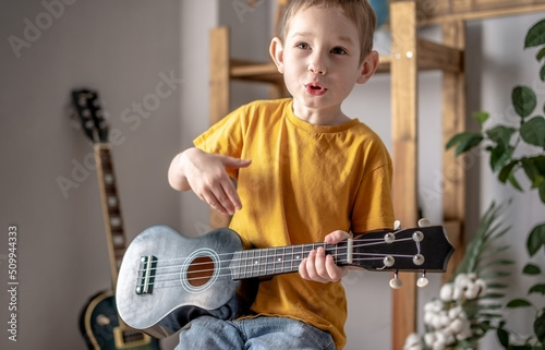 Cute cheerful boy is playing ukulele guitar in the music room. Joyful learning to play musical instruments. photo