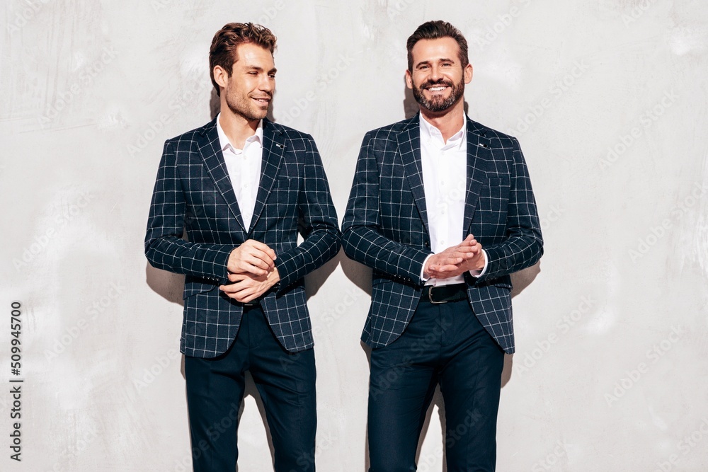 Portrait of two handsome smiling stylish hipster lambersexual models. Sexy modern men dressed in black same elegant suit. Fashion male posing in studio near grey wall. Cheerful and happy Portrait of two handsome smiling stylish hipster lambersexual models. Sexy modern men dressed in black same elegant suit. Fashion male posing in studio near grey wall. Cheerful and happy