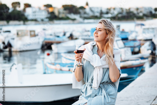 Blond woman with shirt around neck, holding tasty wineglass on vacation in travelling near boat port. Enjoy sunset view © Татьяна Волкова