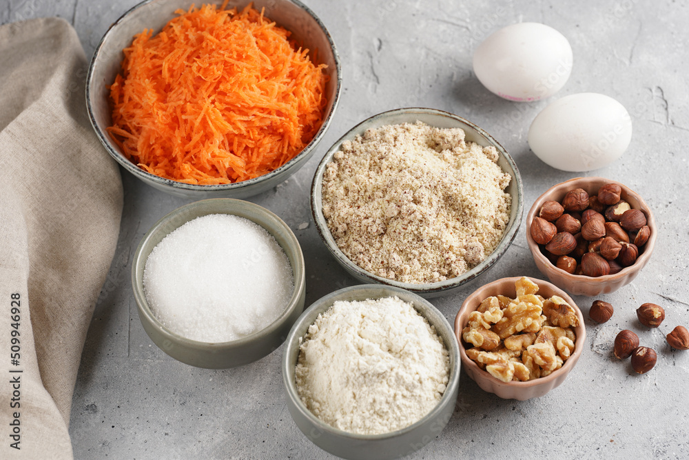 Ingredients for baking a carrot cake: finely grated carrots, grounded hazelnuts and almonds, chopped walnuts, white flour, eggs, sugar on grey background Ingredients for baking a carrot cake: finely grated carrots, grounded hazelnuts and almonds, chopped walnuts, white flour, eggs, sugar on grey background