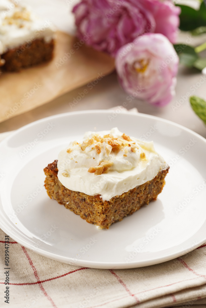 Homemade pastry carrot-walnut cake with grounded almonds and hazelnuts and white cream cheese top layer on white plate on pink checkered napkin