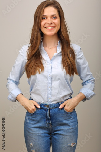 Smiling woman in blue shirt and jeans keeps her hands in pockets. isolated portrait of happy girl on gray.