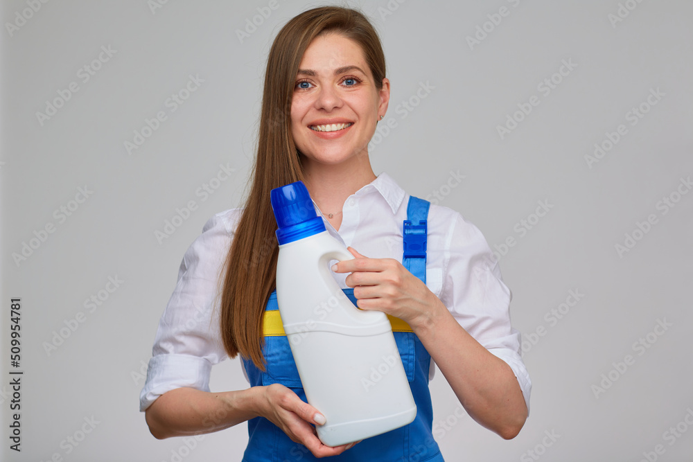 Isolated studio portrait on gray of smiling girl dressed blue overall. Woman ceaning or laundry worker holding big white bottle of detergent.