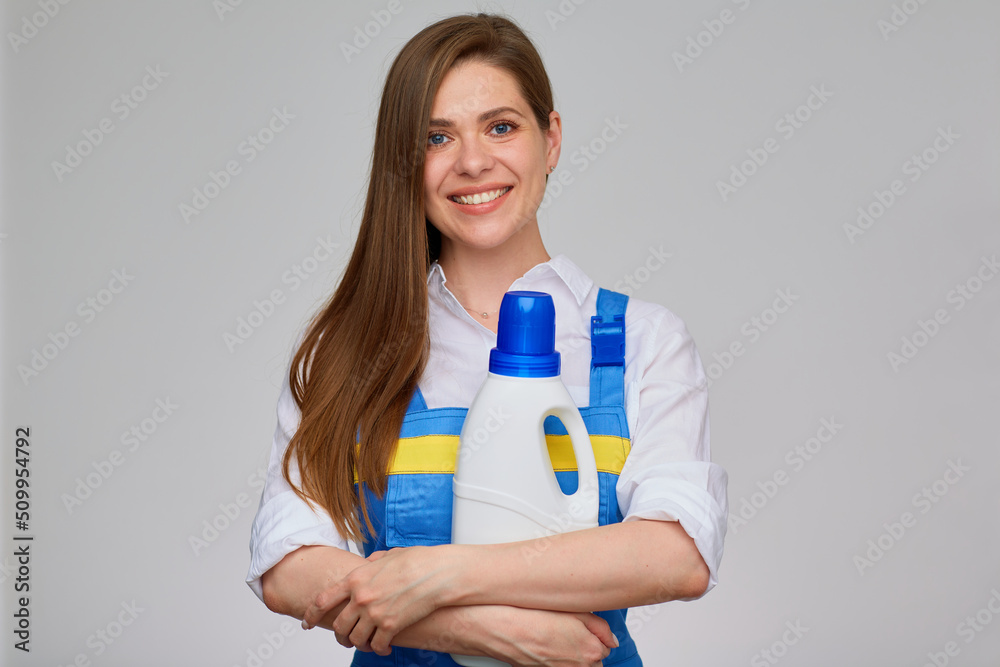Isolated studio portrait on gray of smiling girl dressed blue overall. Woman ceaning or laundry worker holding big white bottle of detergent. Isolated studio portrait on gray of smiling girl dressed blue overall. Woman ceaning or laundry worker holding big white bottle of detergent.