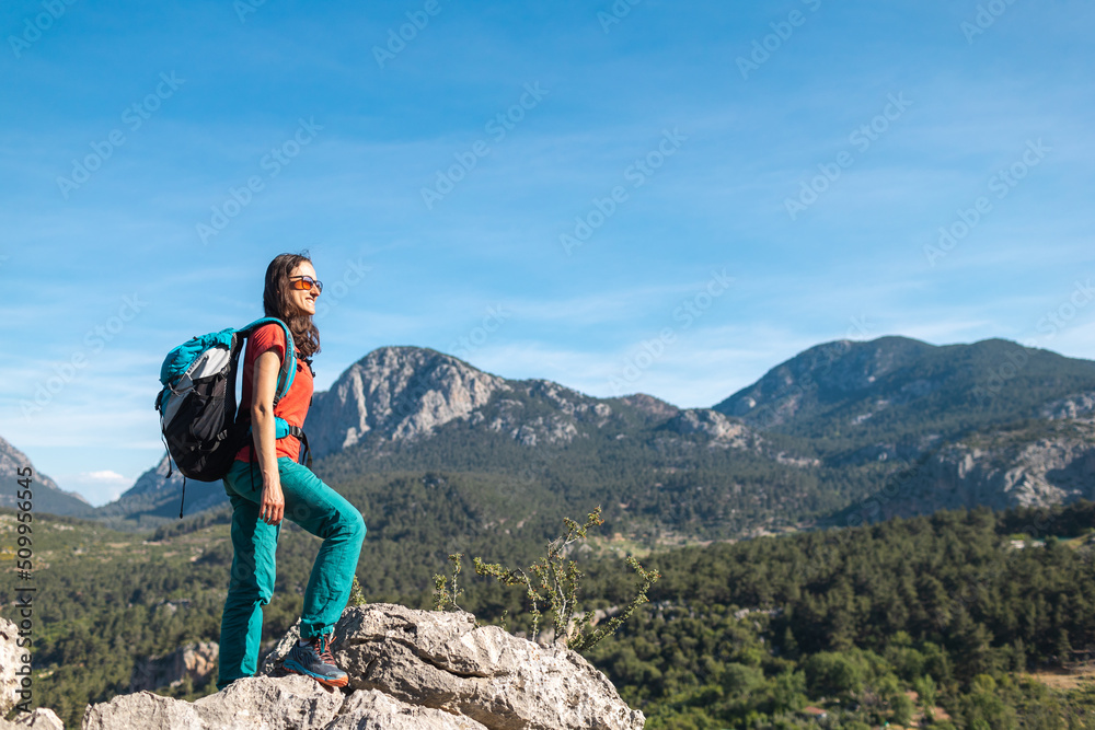 A young woman with a backpack climbed to the top of the mountain and looks at the valley