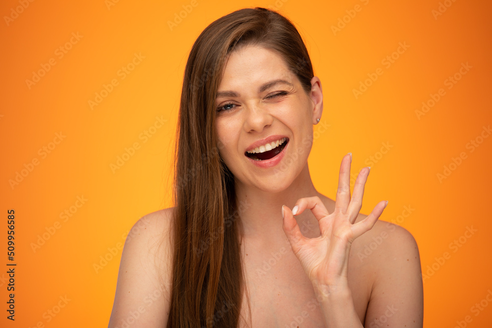 Winking woman with long hair and bare shoulders showing ok gesture. isolated female close up portrait on orange background. Winking woman with long hair and bare shoulders showing ok gesture. isolated female close up portrait on orange background.