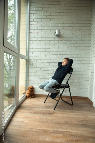 A pensive boy of 6-7 years old sits on a chair on the balcony and looks out the window. Portrait of a boy