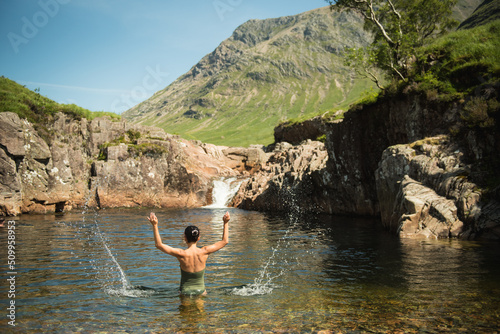 A girl wearing a green swimming suit splashes water with both hands while bathing in the River Etive in Glen Etive, Scottish Highlands, UK, with a waterfall and a mountain in the background.