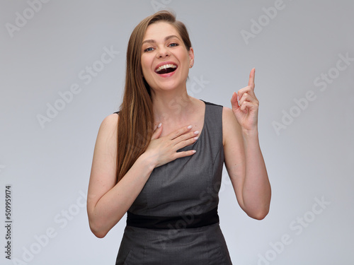 Smiling woman in business dress pointing fingersup and holding one hand on chest, happy girl isolated portrait. photo