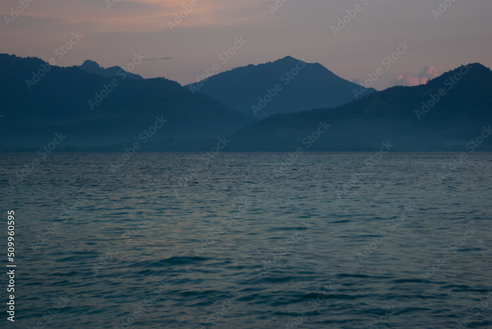 Beautiful early morning on calm sea with blue smooth waves, breeze, pastel pink sky, blue mountains in haze on distant island, horizon. Marine sunrise landscape in indonesian ocean, vacation in asia.