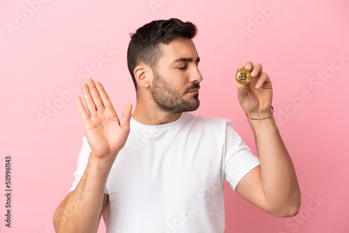 Young man holding a Bitcoin isolated on pink background making stop gesture and disappointed
