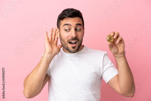 Young man holding a Bitcoin isolated on pink background listening to something by putting hand on the ear