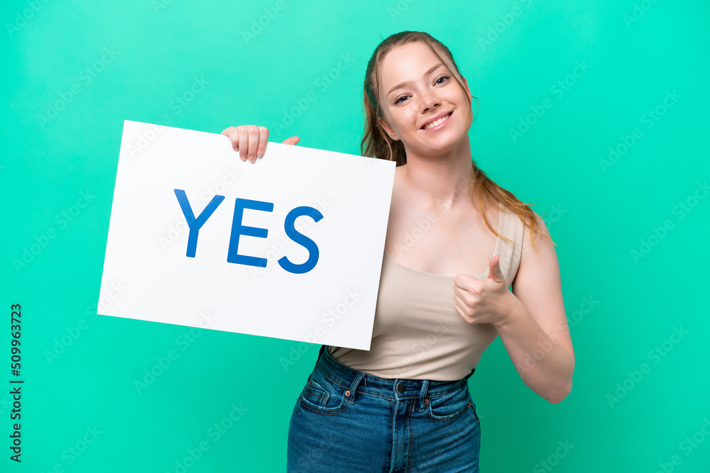 Young caucasian woman isolated on green background holding a placard with text YES with thumb up Young caucasian woman isolated on green background holding a placard with text YES with thumb up