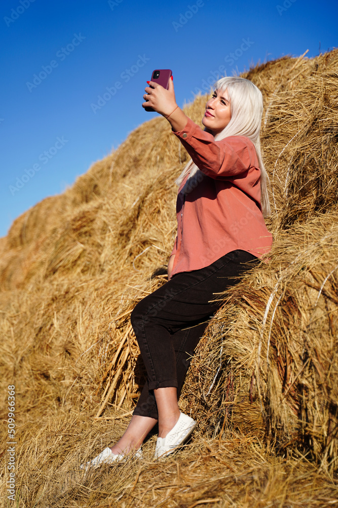 Blonde girl sitting in the hayloft with a phone takes a selfie in autumn