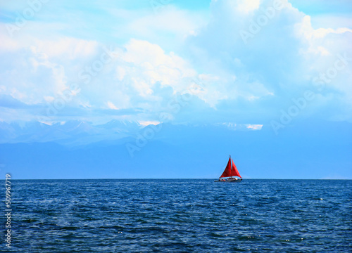 Beautiful romantic view of lonely sailing ship with red sheet against the background of distant mountain range and cloudy blue sky, Issyk-Kul Lake, Kyrgyzstan, Central Asia