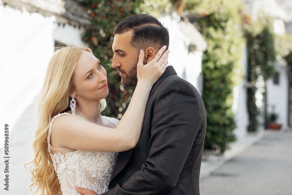 Naklejka premium Portrait of young blonde bride in white dress and brunet groom in suit embracing and looking at each other on city street in Italy, urban background. Beautiful and romantic wedding, happy couple