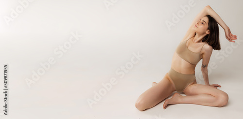 Portrait of beautiful young woman sitting on floor, posing in beige underwear isolated over grey studio background. Sensuality