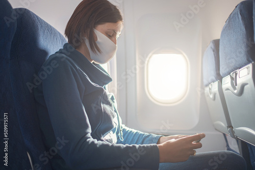Young beautiful woman sitting at window of plane during the flight. new normal travel after covid-19 pandemic concept. reading book on smartphone, playing apps, writing notes