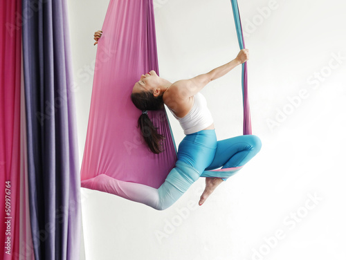 Mature woman pose in hammock performing aerial yoga or flying yoga exercise against white wall background in yoga studio room. Healthy lifestyles and emotional health of middle aged people concepts.