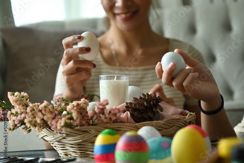 Close up of Asian female holding duck s egg in front of scented candle in a basket over painted multi-colored Easter eggs background.