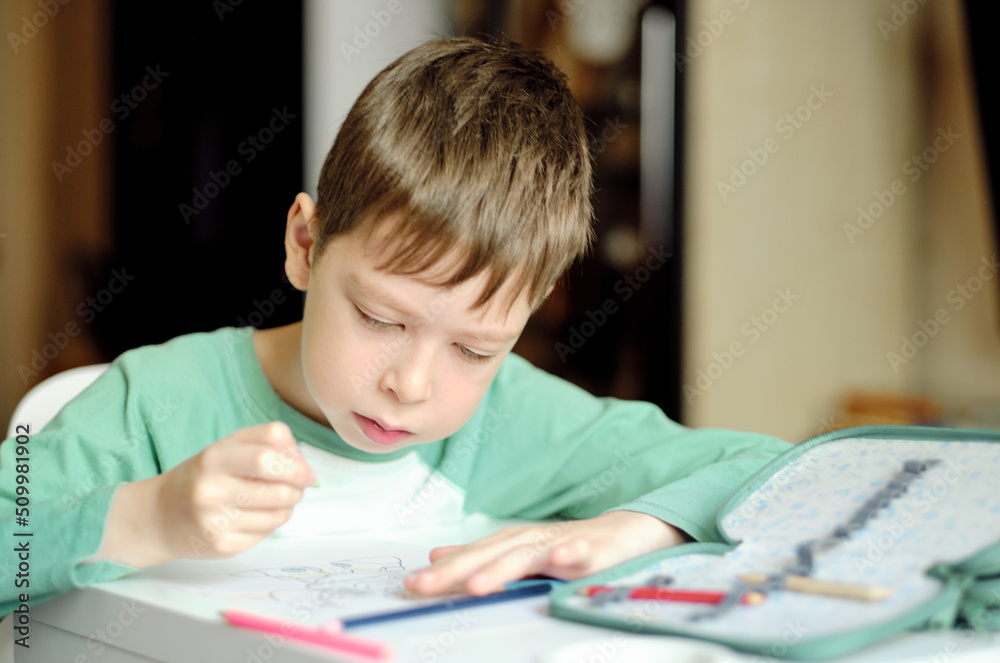 cute and smiling cucasian boy draws with colored pencils while sitting at the table. the boy paints the drawing with a blue pencil. Drawing lessons. Art therapy for stress relief. cute and smiling cucasian boy draws with colored pencils while sitting at the table. the boy paints the drawing with a blue pencil. Drawing lessons. Art therapy for stress relief.