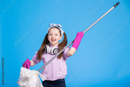 Little girl doing room cleaning, child play cleaning floor with mop and rag at home. Concept of cleanliness and order in the house. Help with housework fun listening to music on blue background.