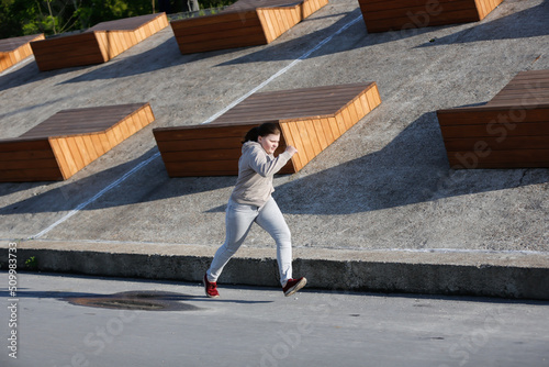 An overweight European teenage girl in hoodie and jeans jogging along concrete embankment, on outskirts of city. Sports and teenagers, overweight teenagers