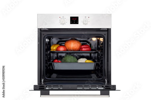 Electric oven with open door, lights on and vegetables for roasting. Front view. Isolated on white