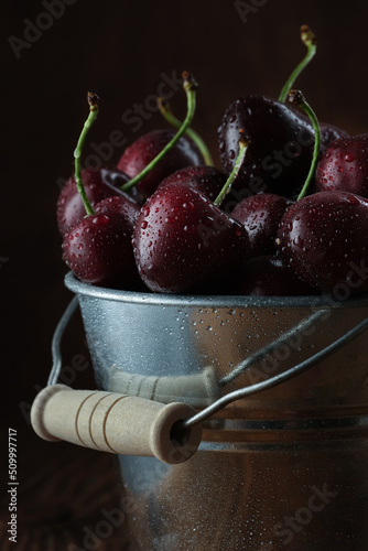 Ripe dark red cherries with water drops in a metal bucket on a wooden background. Theme of healthy food.