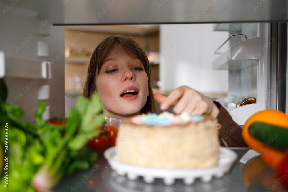 Young woman tasting cake with finger in fridge