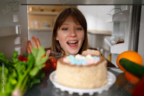 Young woman looking at delicious cake in fridge