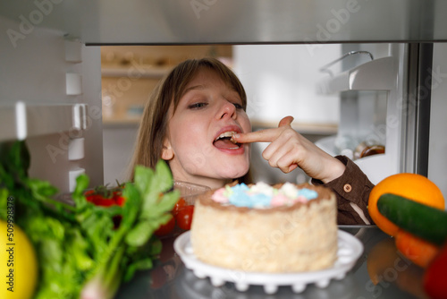 Cheerful woman tasting cake with finger in fridge